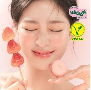 Advert for Korean lip gloss, showing a smiling woman with her eyes closed who's wearing the product. She's holding the packaged product in one hand and strawberries on a stick in the other hand.